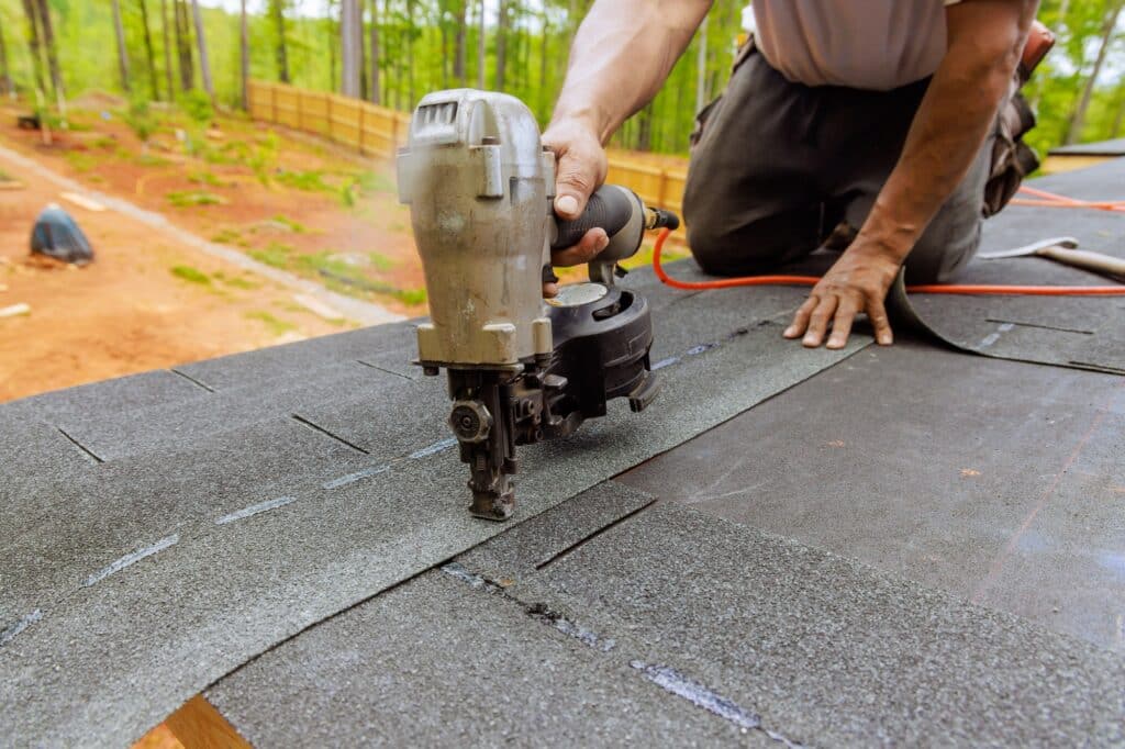 Using an air pneumatic nail gun, a roofer install new asphalt bitumen shingles